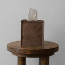 Brown leather tissue holder with tissues on a wooden table against a plain background