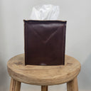 Brown leather tissue holder with a white tissue on a wooden stool against a gray background