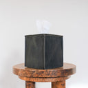 A dark charcoal gray leather tissue box cover with visible grain texture and white tissue paper partially pulled out, displayed on a rustic round wooden stool against a plain white background.