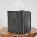 A square black leather tissue box cover with visible stitching and a white tissue protruding from the top, displayed on a round wooden surface against a plain light background.