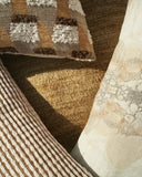 Close-up of textured home decor textiles, including a woven throw pillow with a geometric pattern in brown, beige, and white, a striped pillow, and a sheer patterned fabric panel on a warm brown background.