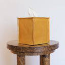 Mustard-colored leather tissue box cover on a wooden pedestal against a white background