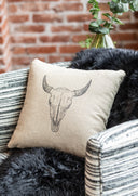 Cream throw pillow with embroidered longhorn skull design, styled on a dark fur throw against a rustic brick wall background.