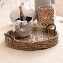 A round woven seagrass serving tray with dark metal handles, holding a silver shell-shaped wine bottle cooler with a bottle and two clear glasses, set on a light wood table.