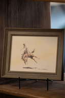 Framed art print featuring a bucking bronco, framed in rustic wood and displayed on an easel on wood shelving, 