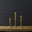 Three brass candlesticks of different heights displayed on a rustic wooden table against a dark gray wood-paneled wall.