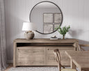 Wooden sideboard with a lamp and decorative items in a room with a round mirror and wall art.