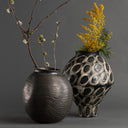 Two black ceramic vases with textured surfaces, one containing branches and the other with yellow flowers, on a dark background.