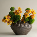 A decorative floral arrangement featuring bright yellow orchids and green moss balls in a textured dark gray ceramic pot, photographed against a plain light gray studio background.