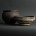 Two rustic handmade clay vessels, a large rounded pot and a wide shallow bowl, shown on a dark gray studio background.