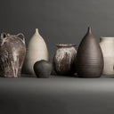 A group of seven handmade ceramic vases in various shapes and earthy tones, including dark brown, charcoal gray, beige, and white marbled textures, arranged on a dark gray surface against a matching dark gray background.
