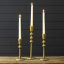Three gold-toned metal candlestick holders with white taper candles, arranged on a rustic wooden surface against a dark charcoal paneled background.