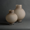 Two round glass vases with short necks and clear glass bases, shown on a dark gray studio background. The vases have a warm taupe-beige color with subtle brown shading near the rims and clear glass at the base.