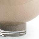Close-up of a large, smooth, rounded vessel or planter in a warm beige or blush-tinted tone, with a thick clear glass base, shown against a white background.