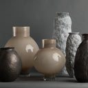 A collection of modern decorative vases in various shapes and textures, including glossy beige globes, textured gray stone-like forms, and dark metallic ribbed vases, arranged on a dark gray surface against a matching gray background.