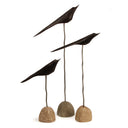 Set of three black wood and metal bird sculptures on slender stands with natural stone bases with white background