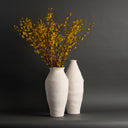 Two textured white ceramic vases on a dark gray surface against a dark gray background, with yellow flowering branches arranged in the taller vase.