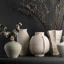 A collection of five textured ceramic vases in off-white and light gray, styled with cotton bolls and willow branches against a dark gray background.
