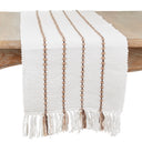 A white woven table runner with vertical tan stripes and fringe tassels, draped over a wooden table against a white background.