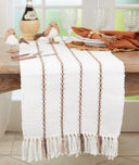 A white woven table runner with vertical tan stripes and fringe tassels, styled on a wooden dining table with place settings, glassware, decorative birds, and a bottle in a bright room.