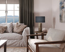 A styled living room corner featuring a light beige slipcovered sofa with neutral pillows, a matching armchair, a dark gray curtain, and a table lamp with a textured gold base on a wooden side table.