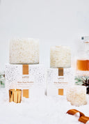 Two different sizes of the LAFCO White Maple Bourbon scented candle in a white speckled glass vessel with warm amber accents sitting on a white and gold confetti boxes  