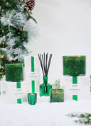 LAFCO Woodland Spruce scented candles and a diffuser in vibrant green glass vessels with subtle gold marbling in different sizes. Sitting on top of the white, green confetti box with evergreen & snow accents