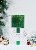 LAFCO Woodland Spruce scented candle in a vibrant green glass vessel with subtle gold marbling.  Sitting on top of the white, green confetti box with evergreen, pinecone & snow accents