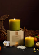 A LAFCO 'Pistachio Nut' scented candle in a clear glass jar with a yellow-green wax, styled on top of its beige cardboard packaging box. A second, unlit candle in the same jar sits to the right, and the scene is decorated with dried leaves, a white orchid, and pistachios against a dark background.
