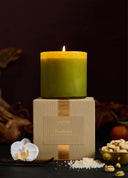 A lit green candle displayed on top of a beige square box with a gold ribbon, set against a dark background with decorative botanical and food props.