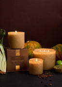 Three lit Lafoi candles in textured beige holders are arranged with a brown branded candle box and autumn ingredients including a fennel bulb, pumpkin, and green lime on a dark surface against a dark brown background.