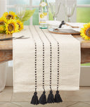 An off-white woven table runner with black vertical dotted stripes and four black tassels at the bottom, styled on a wooden dining table with sunflowers, wine glasses, and a bottle of sparkling water.