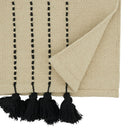 A close-up of a woven throw blanket in a warm cream or beige color, featuring vertical rows of black woven stripes and black tassels along one edge, with one corner folded over to show the texture.