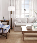A bright, modern living room featuring a light beige sofa, a blue and white patterned armchair, a wooden coffee table, and a floor lamp in front of a large window with sheer curtains.