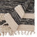A close-up of a woven throw blanket with a black and cream striped pattern, featuring chunky cream pom-pom trim and fringe tassels.