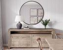 Wooden sideboard with round mirror, Theo Table lamp, and decor items against a light wooden paneled wall.