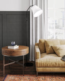 A modern living room scene featuring a round wooden side table with a glass top and metal legs, next to a beige velvet sofa and a white floor lamp.