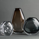 Three modern glass vases in different shapes and colors: a clear bubble-textured glass sphere, a tall translucent smoky brown vase, and a dark charcoal glass bowl, arranged on a neutral gray background.